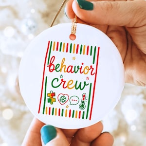 May include: A round white ceramic ornament featuring the text behavior crew in red and green cursive script. The design includes a colorful puzzle piece, a heart with a snowflake, a speech bubble, and a thermometer icon, framed by vertical stripes.