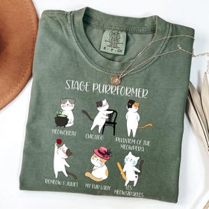 May include: Green t-shirt with the text "STAGE PURRFORMER" and illustrations of cats in theatrical roles. The shirt features the Comfort Colors brand label and a gold necklace. The cat illustrations include "Meowcbeth," "Chicatgo," and "Phantom of the Meowpera."