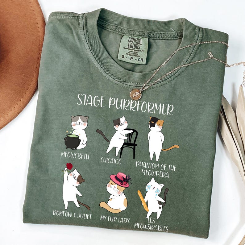 May include: Green t-shirt with the text "STAGE PURRFORMER" and illustrations of cats in theatrical roles. The shirt features the Comfort Colors brand label and a gold necklace. The cat illustrations include "Meowcbeth," "Chicatgo," and "Phantom of the Meowpera."
