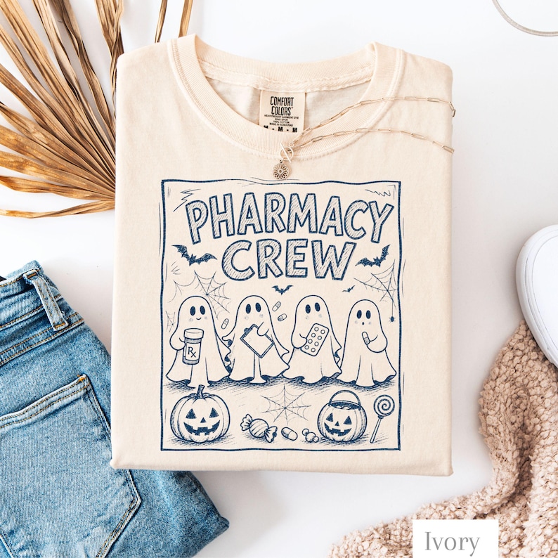 K&ouml;nnte beinhalten: Ein elfenbeinfarbenes T-Shirt mit einem dunkelblauen Grafikdruck und dem Text PHARMACY CREW. Das Design zeigt vier Geister mit Apothekenartikeln, einen K&uuml;rbis, einen S&uuml;&szlig;igkeiten-Eimer, Spinnweben, Flederm&auml;use und verschiedene Pillen im gesamten Motiv.