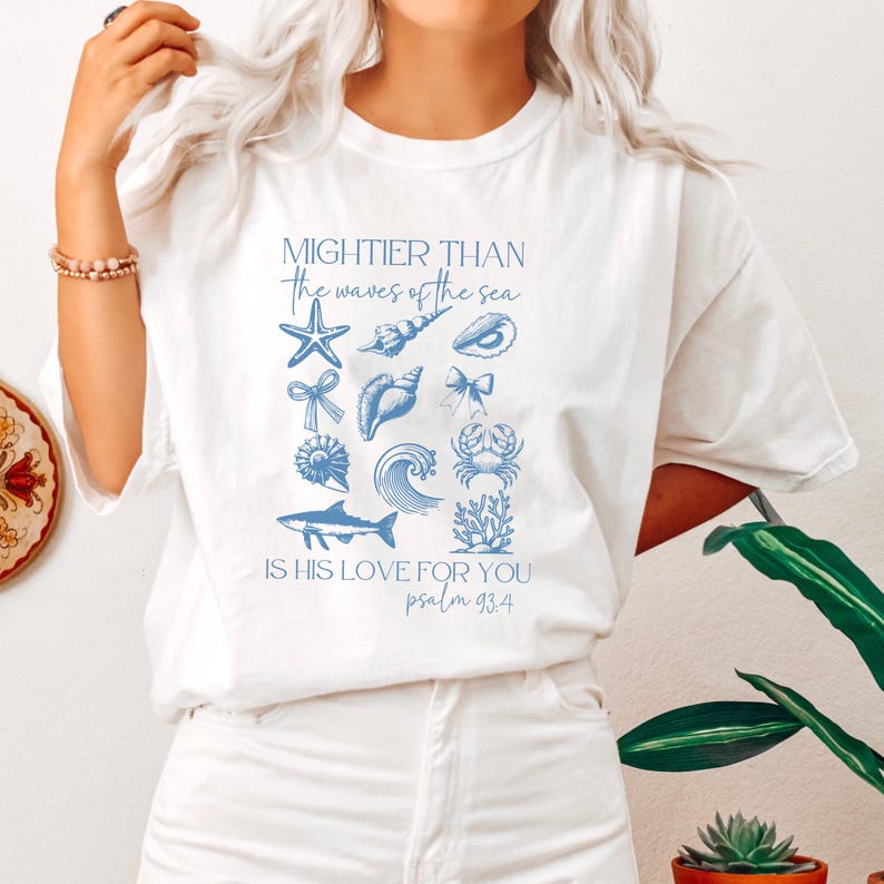 May include: White t-shirt with blue sea-themed graphics and text. The design includes a starfish, seashells, bows, a wave, a shark, and coral. The text reads "Mightier than the waves of the sea, is his love for you, psalm 93:4."