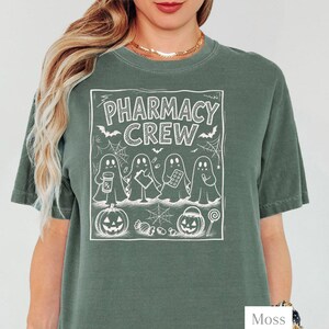 K&ouml;nnte beinhalten: Ein moosgr&uuml;nes Kurzarm-T-Shirt mit einem wei&szlig;en Grafikdruck von vier Geistern und dem Text PHARMACY CREW. Das Design enth&auml;lt Illustrationen einer Medikamentendose, eines Klemmbretts, einer Pillenpackung, eines K&uuml;rbisses und eines Lollis.