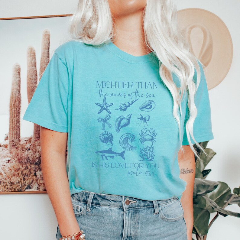 May include: A light blue t-shirt featuring the text "MIGHTIER THAN the waves of the sea" and "IS HIS LOVE FOR YOU psalm 93:4". The shirt has a graphic of various sea creatures in a light blue color.
