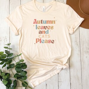 K&ouml;nnte beinhalten: Ein beiges T-Shirt mit dem Text "Autumn leaves and cats please" in bunten, stilisierten Buchstaben.