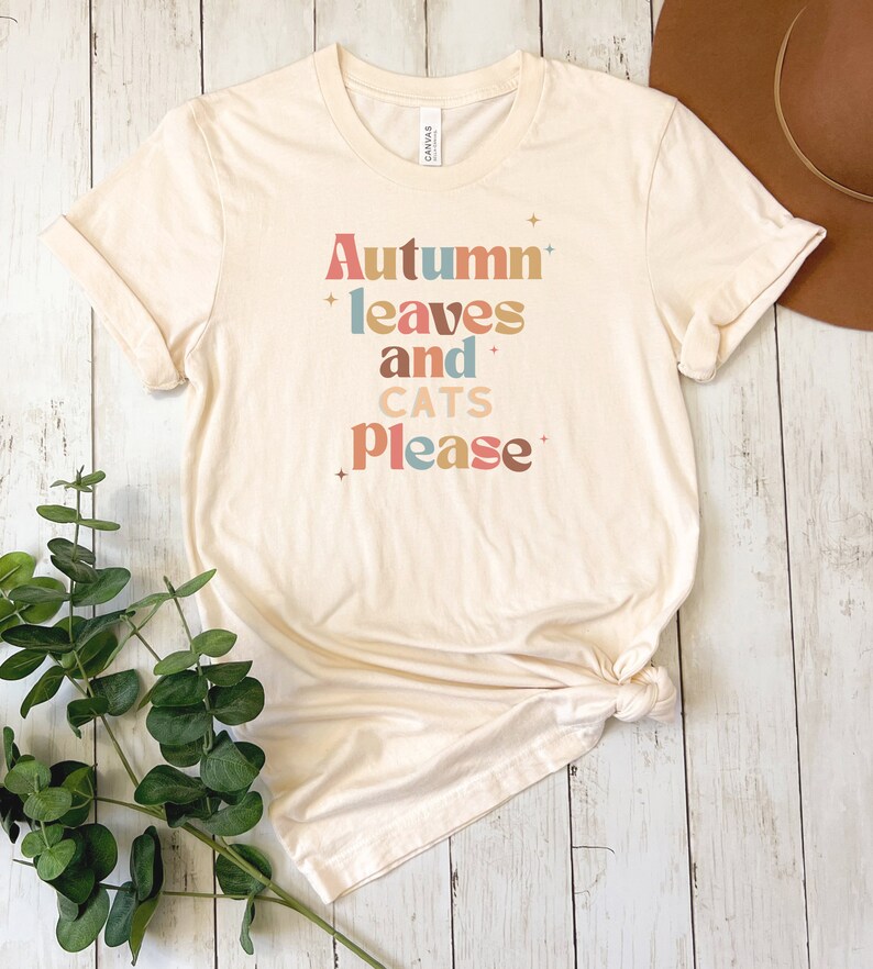 K&ouml;nnte beinhalten: Ein beiges T-Shirt mit dem Text "Autumn leaves and cats please" in bunten, stilisierten Buchstaben.