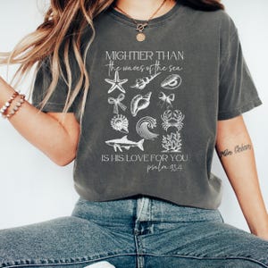 May include: Dark gray t-shirt with white illustrations of sea creatures and the text "MIGHTIER THAN the waves of the sea IS HIS LOVE FOR YOU psalm 93.4". The shirt is a comfortable, casual style.