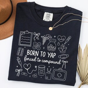 May include: Navy blue t-shirt with white medical-themed illustrations and the text "BORN TO YAP forced to compound." The shirt features a crew neck and short sleeves. A gold necklace and a brown hat are also visible.