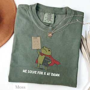 May include: An olive green t-shirt featuring a graphic of a frog wearing a red cape, holding a book titled Graphing and a sign labelled Algebra with maths equations. The text below reads We Solve For X At Dawn. The shirt has a Comfort Colors label.