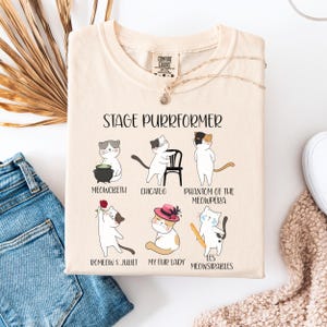 May include: Cream-colored t-shirt with the text "STAGE PURRFORMER" and illustrations of cats in theatrical roles. The cats are depicted as characters from famous plays, including "Meowcbeth" and "Romeo & Juliet".