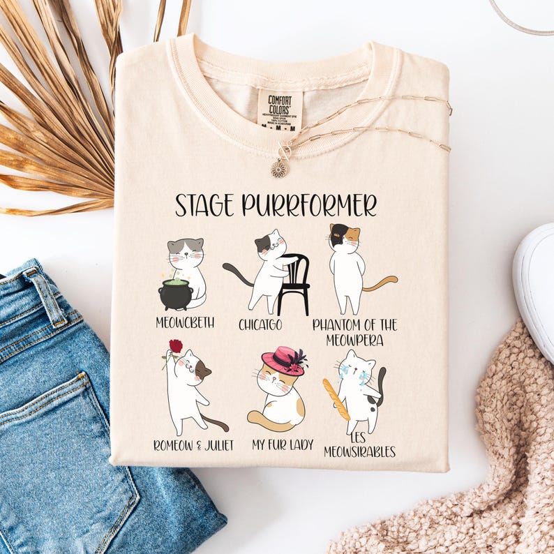 May include: Cream-colored t-shirt with the text "STAGE PURRFORMER" and illustrations of cats in theatrical roles. The cats are depicted as characters from famous plays, including "Meowcbeth" and "Romeo & Juliet".