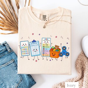 May include: An ivory t-shirt featuring a graphic print of anthropomorphic items including a pop-it toy, an ABA data sheet clipboard, an ABA visual schedule, a toy bin with a teddy bear, and a blue fidget spinner, all with smiling faces.