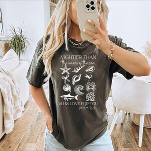 May include: Dark gray t-shirt featuring a collection of white sea-themed illustrations, including starfish, shells, and a wave. The text reads "Mightier than the waves of the sea, is his love for you, psalm 93:4."