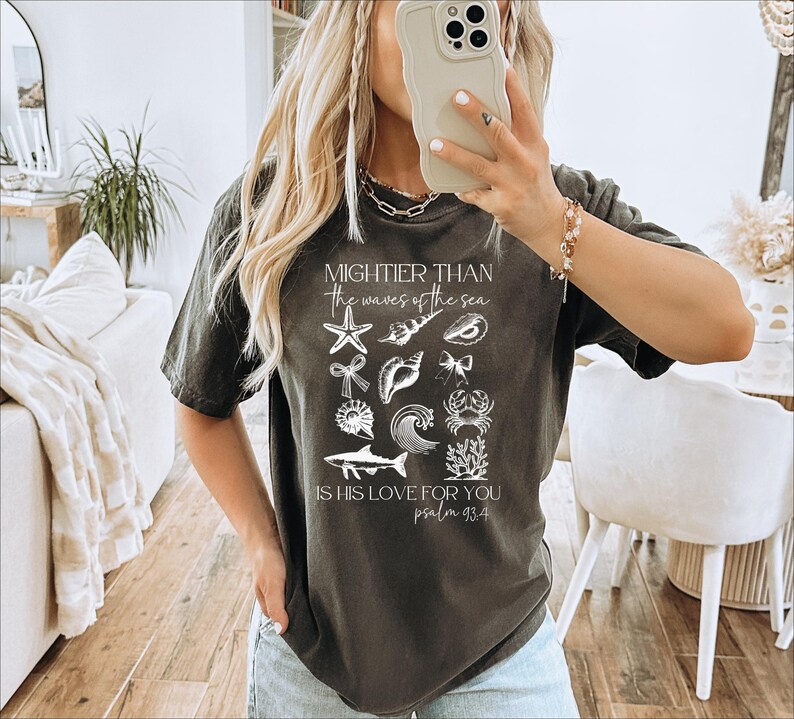 May include: Dark gray t-shirt featuring a collection of white sea-themed illustrations, including starfish, shells, and a wave. The text reads "Mightier than the waves of the sea, is his love for you, psalm 93:4."