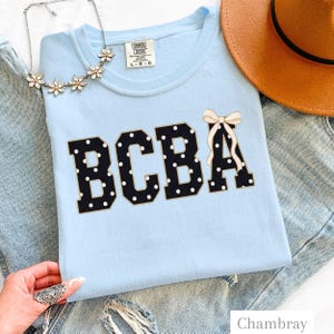 Könnte beinhalten: Ein hellblaues T-Shirt mit den Buchstaben BCBA in Schwarz mit weißen Punkten und einer cremefarbenen Schleifengrafik auf dem Buchstaben A. Das T-Shirt hat ein Comfort Colors-Etikett am Kragen und wird mit einer silbernen Blumenkette und einem beigen Hut gezeigt.