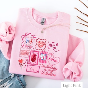 Könnte beinhalten: Hellrosa Sweatshirt mit einem grafischen Design mit Herzen, anatomischen Diagrammen und dem Text "Cardiology is my Valentine". Das Design beinhaltet ein Stethoskop und einen herzförmigen Lutscher. Das Sweatshirt ist aus weichem Material.