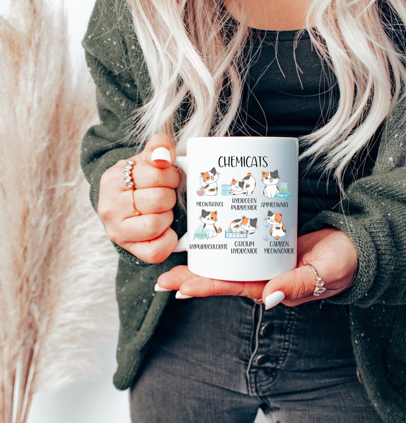 K&ouml;nnte beinhalten: Eine wei&szlig;e Keramiktasse mit einer humorvollen Illustration von Katzen, die verschiedene chemische Verbindungen darstellen. Der Text "Chemicats" ist oben auf der Tasse aufgedruckt. Die Katzenillustrationen sind mit den Namen der chemischen Verbindungen beschriftet, wie z. B. "Methanol", "Wasserstoffperoxid" und "Calciumhydroxid".