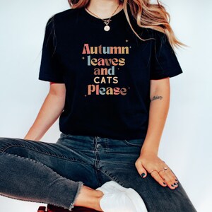 K&ouml;nnte beinhalten: Ein schwarzes T-Shirt mit dem Text "Autumn leaves and cats please" in bunten, Retro-Lettern.