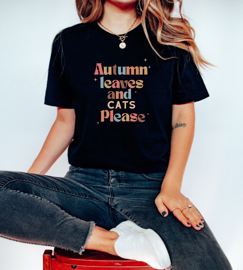 K&ouml;nnte beinhalten: Ein schwarzes T-Shirt mit dem Text "Autumn leaves and cats please" in bunten, Retro-Lettern.
