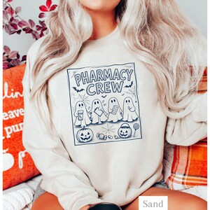 K&ouml;nnte beinhalten: Ein sandfarbenes Sweatshirt mit Rundhalsausschnitt und einem dunkelblauen Grafikdruck. Das Design zeigt vier Geister, zwei K&uuml;rbisse, Spinnweben und S&uuml;&szlig;igkeiten, mit dem Text PHARMACY CREW &uuml;ber den Geistern, die medizinische Utensilien halten.