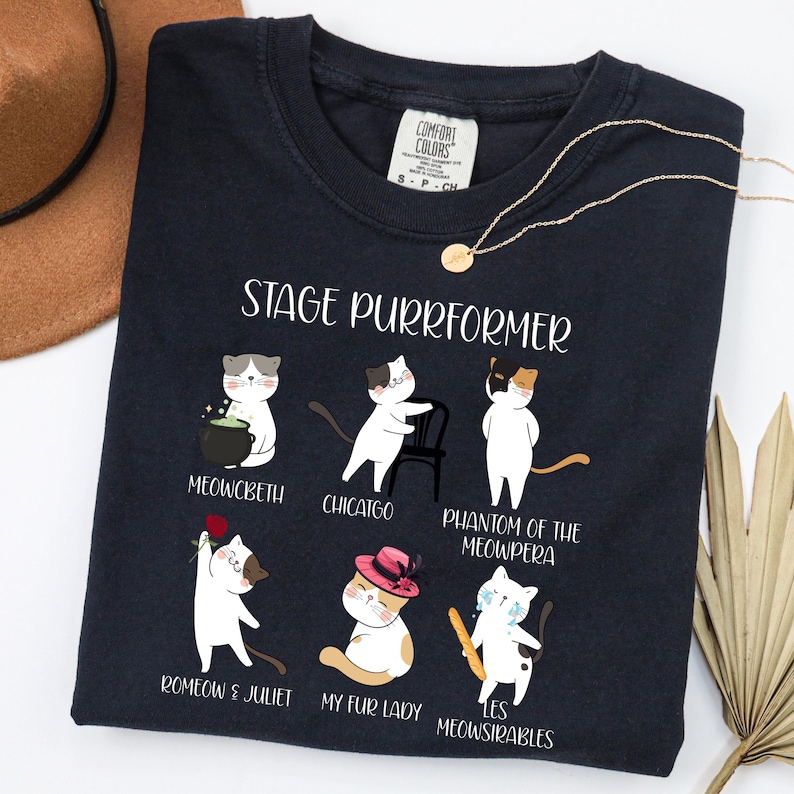 May include: Navy blue t-shirt with the text "STAGE PURRFORMER" and illustrations of cats in theatrical roles. The shirt features cats in roles such as "Meowcbeth" and "Romeow & Juliet". A gold necklace is draped on the shirt.