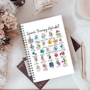 May include: A white spiral-bound notebook titled "Speech Therapy Alphabet". The cover displays an alphabet with speech therapy terms like "dysphagia" and "intonation", each paired with a small illustration. The notebook is on a desk.