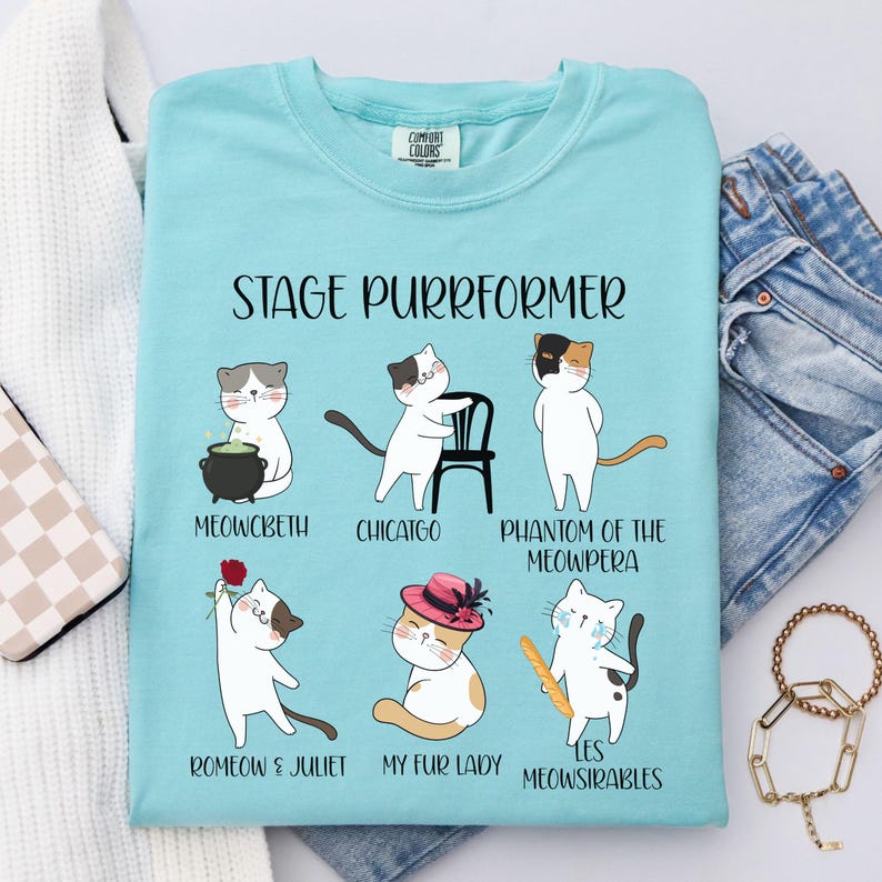 May include: Light blue t-shirt with the text "STAGE PURRFORMER" and illustrations of cats in theatrical roles. The cats are depicted as characters from famous plays and musicals, including "Meowcbeth" and "Chicatgo".