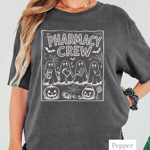 K&ouml;nnte beinhalten: Ein anthrazitfarbenes Kurzarm-T-Shirt mit einem wei&szlig;en Grafikdruck von vier Geistern, die Apothekenartikel halten. Der Text lautet PHARMACY CREW, erg&auml;nzt durch Illustrationen von Flederm&auml;usen, Spinnennetzen, zwei K&uuml;rbissen und verschiedenen S&uuml;&szlig;igkeiten.