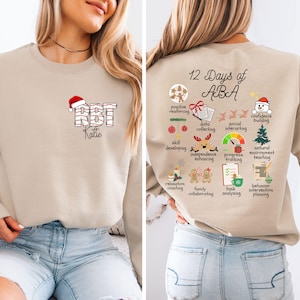 Pode incluir: Camisola bege com o texto "RBT Katie" num design festivo na frente. A parte de trás apresenta "12 Days of ABA" com ícones relacionados. Uma peça de vestuário confortável e educativa.