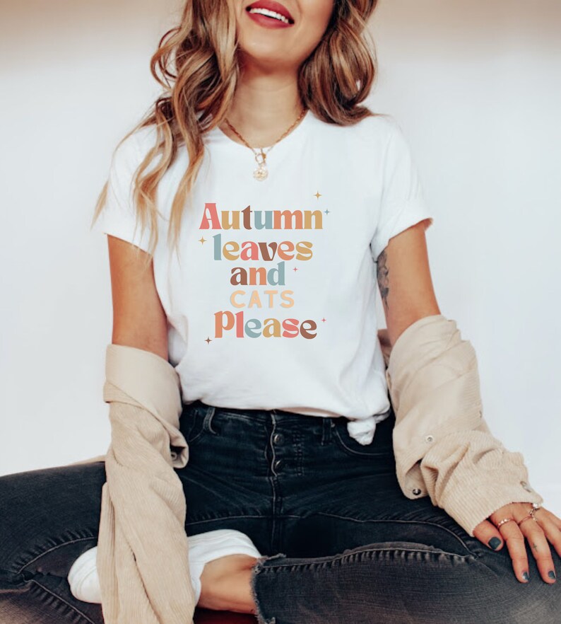 K&ouml;nnte beinhalten: Ein wei&szlig;es T-Shirt mit dem Text "Autumn leaves and cats please" in bunten, Retro-Lettern.