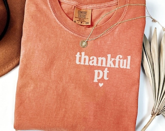 Può includere: Una t-shirt color terracotta con scollo rotondo e un'etichetta del marchio Comfort Colors sul colletto interno. La parte anteriore presenta la scritta bianca "thankful pt" con una piccola icona a forma di cuore sotto le lettere. Una collana dorata poggia sulla t-shirt.