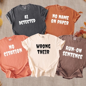 May include: A collection of five short-sleeve crew neck t-shirts in charcoal, orange, peach, cream, and brown. The shirts feature white or black text prints reading AI DETECTED, NO NAME ON PAPER, NO CITATION, WRONG THEIR, and RUN-ON SENTENCE.
