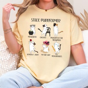 May include: Yellow t-shirt with the text "STAGE PURRFORMER" and illustrations of cats in theatrical roles. The cats are depicted in scenes from "Meowcbeth," "Chicago," "Phantom of the Meowpera," "Romeow & Juliet," "My Fur Lady," and "Les Meowsirables."