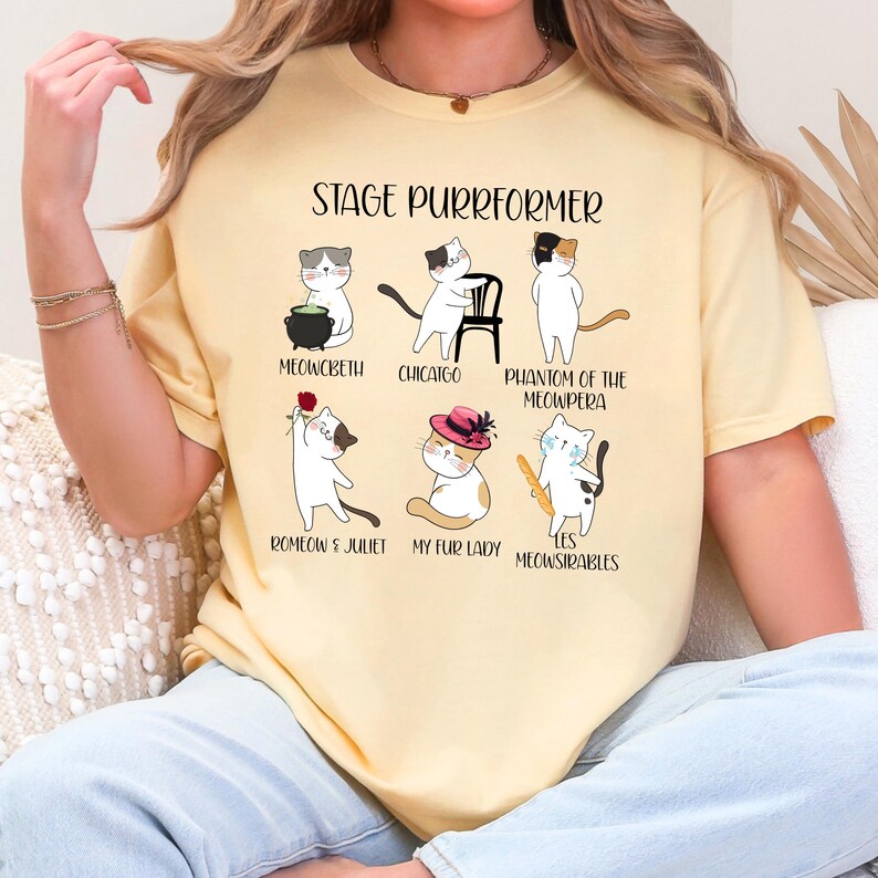 May include: Yellow t-shirt with the text "STAGE PURRFORMER" and illustrations of cats in theatrical roles. The cats are depicted in scenes from "Meowcbeth," "Chicago," "Phantom of the Meowpera," "Romeow & Juliet," "My Fur Lady," and "Les Meowsirables."
