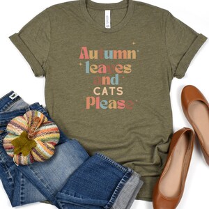 K&ouml;nnte beinhalten: Ein gr&uuml;nes T-Shirt mit dem Text "Autumn leaves and cats please" in bunten Buchstaben. Das Shirt ist gefaltet und liegt auf einer wei&szlig;en Oberfl&auml;che mit einem Paar braunen Ballerinas und einer blauen Jeans mit einem gestrickten K&uuml;rbis.