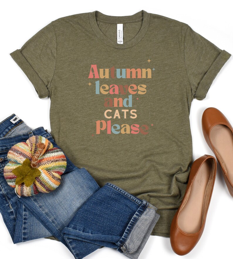 K&ouml;nnte beinhalten: Ein gr&uuml;nes T-Shirt mit dem Text "Autumn leaves and cats please" in bunten Buchstaben. Das Shirt ist gefaltet und liegt auf einer wei&szlig;en Oberfl&auml;che mit einem Paar braunen Ballerinas und einer blauen Jeans mit einem gestrickten K&uuml;rbis.