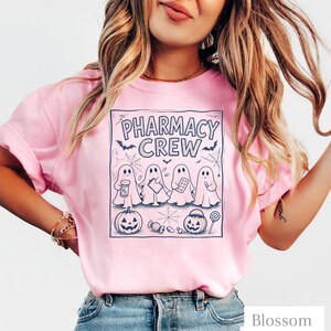 K&ouml;nnte beinhalten: Ein hellrosa T-Shirt mit einem dunkelblauen Grafikdruck und dem Text PHARMACY CREW. Das Design zeigt vier Geister, die Apothekenartikel halten, zwei K&uuml;rbisse, Spinnweben, Flederm&auml;use und verschiedene S&uuml;&szlig;igkeiten in einem rechteckigen Rahmen.