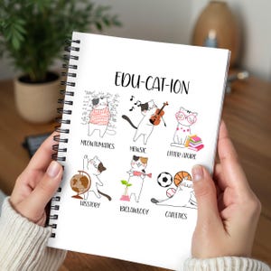 May include: A spiral-bound notebook with the title "EDU-CAT-ION" and illustrations of cats representing different subjects like math, music, and history. The notebook is held by a person wearing a white sweater.