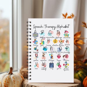 May include: A spiral-bound notebook titled "Speech Therapy Alphabet" featuring the alphabet with speech therapy terms and illustrations. The open notebook displays colourful, hand-drawn images and text, ideal for educational purposes. The notebook is on a wooden surface.