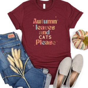 K&ouml;nnte beinhalten: Ein maroonfarbenes T-Shirt mit dem Text "Autumn leaves and cats please" in einer bunten Retro-Schrift.