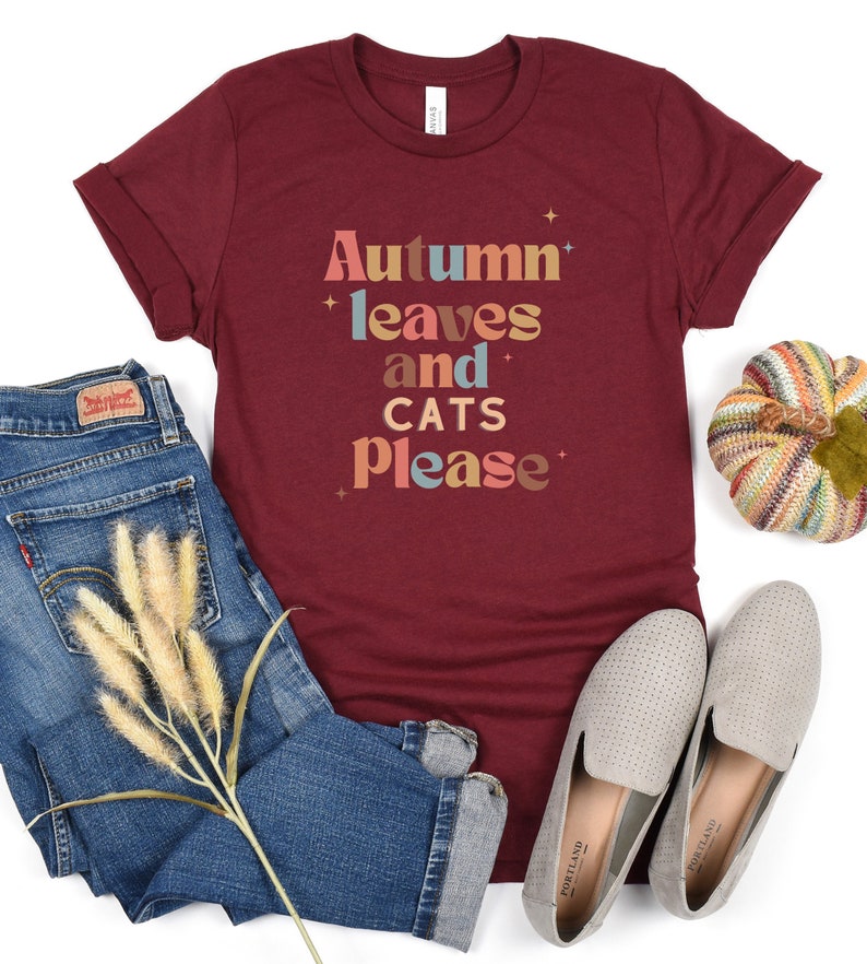 K&ouml;nnte beinhalten: Ein maroonfarbenes T-Shirt mit dem Text "Autumn leaves and cats please" in einer bunten Retro-Schrift.
