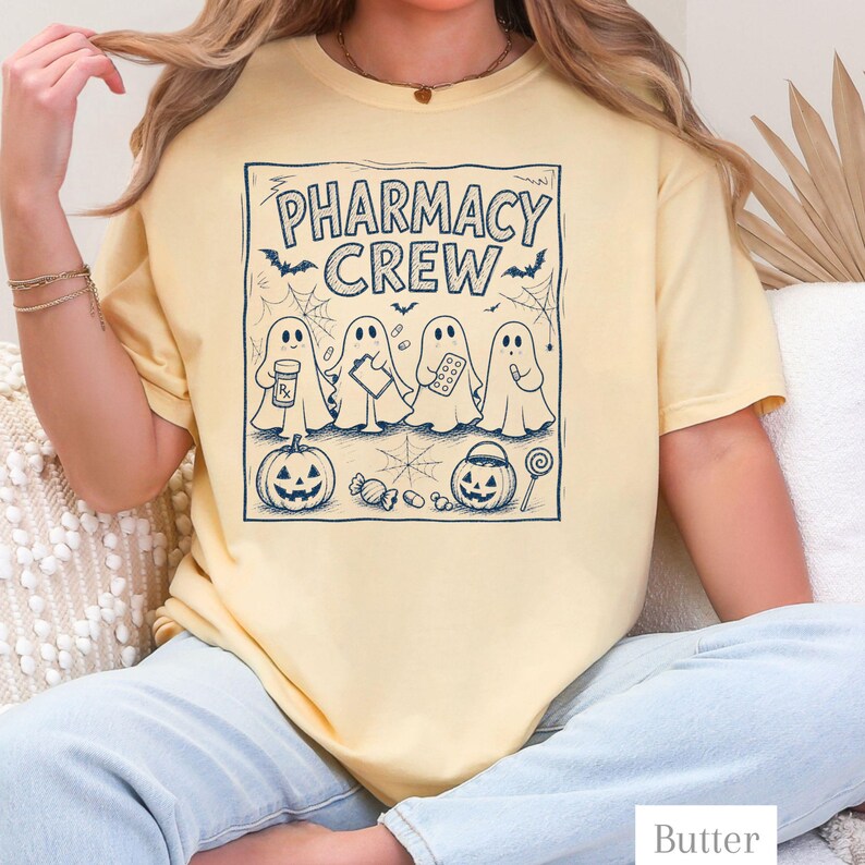 K&ouml;nnte beinhalten: Ein blassgelbes T-Shirt mit einem dunkelblauen Grafikdruck und dem Text PHARMACY CREW. Das Design zeigt vier Geister, die Apothekenartikel halten, umgeben von Flederm&auml;usen, Spinnenweben, zwei K&uuml;rbissen, S&uuml;&szlig;igkeiten und einem Lutscher.
