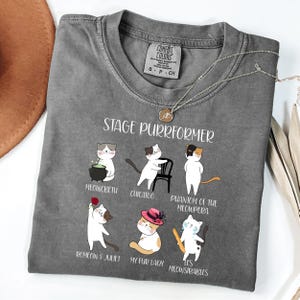 May include: Gray Comfort Colors t-shirt with the text "STAGE PURRFORMER" and illustrations of cats in theatrical roles. The shirt features a crew neck and short sleeves. A gold necklace is draped across the shirt.