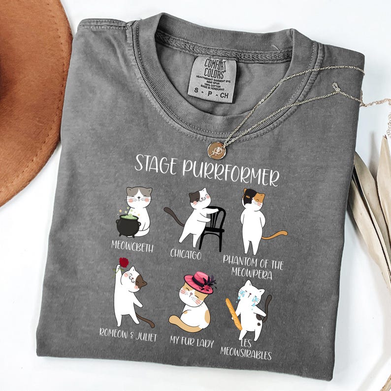 May include: Gray Comfort Colors t-shirt with the text "STAGE PURRFORMER" and illustrations of cats in theatrical roles. The shirt features a crew neck and short sleeves. A gold necklace is draped across the shirt.