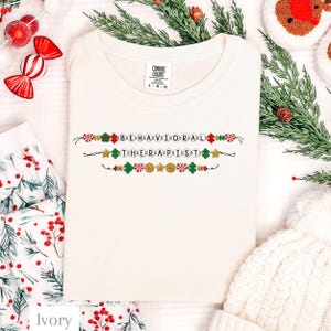 May include: An ivory t-shirt featuring a graphic design of three horizontal strings of beads. The beads spell out the text BEHAVIORAL THERAPIST and are interspersed with red and green puzzle pieces, gold stars, and red and white peppermint candies.