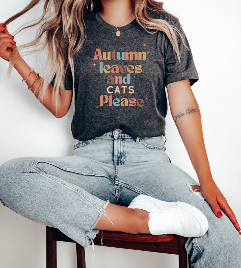 K&ouml;nnte beinhalten: Ein dunkelgraues T-Shirt mit dem Text "Autumn leaves and cats please" in bunten Buchstaben.