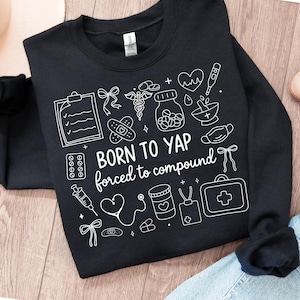 May include: Black sweatshirt featuring white medical illustrations and the text "BORN TO YAP forced to compound." The design includes a clipboard, pills, a syringe, a stethoscope, and a first aid kit.