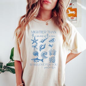 May include: Cream-colored t-shirt featuring blue illustrations of seashells, a starfish, and a wave, with the text "Mightier than the waves of the sea" and "Is His love for you Psalm 93:4".