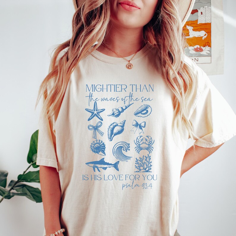 May include: Cream-colored t-shirt featuring blue illustrations of seashells, a starfish, and a wave, with the text "Mightier than the waves of the sea" and "Is His love for you Psalm 93:4".