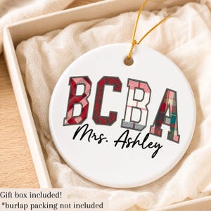 May include: A round white ceramic ornament featuring the letters BCBA in a varsity font with a red and green plaid pattern. Below the letters, the name Mrs. Ashley is printed in a black cursive script. A gold string is attached to the top.