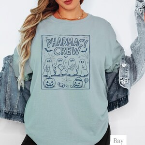 K&ouml;nnte beinhalten: Ein hellblaues Sweatshirt mit Rundhalsausschnitt und einem dunkelblauen Grafikdruck mit dem Text PHARMACY CREW. Das Design zeigt vier Geister, zwei K&uuml;rbisse, Spinnweben, Flederm&auml;use und verschiedene S&uuml;&szlig;igkeiten. Es wird unter einer Jeansjacke getragen.